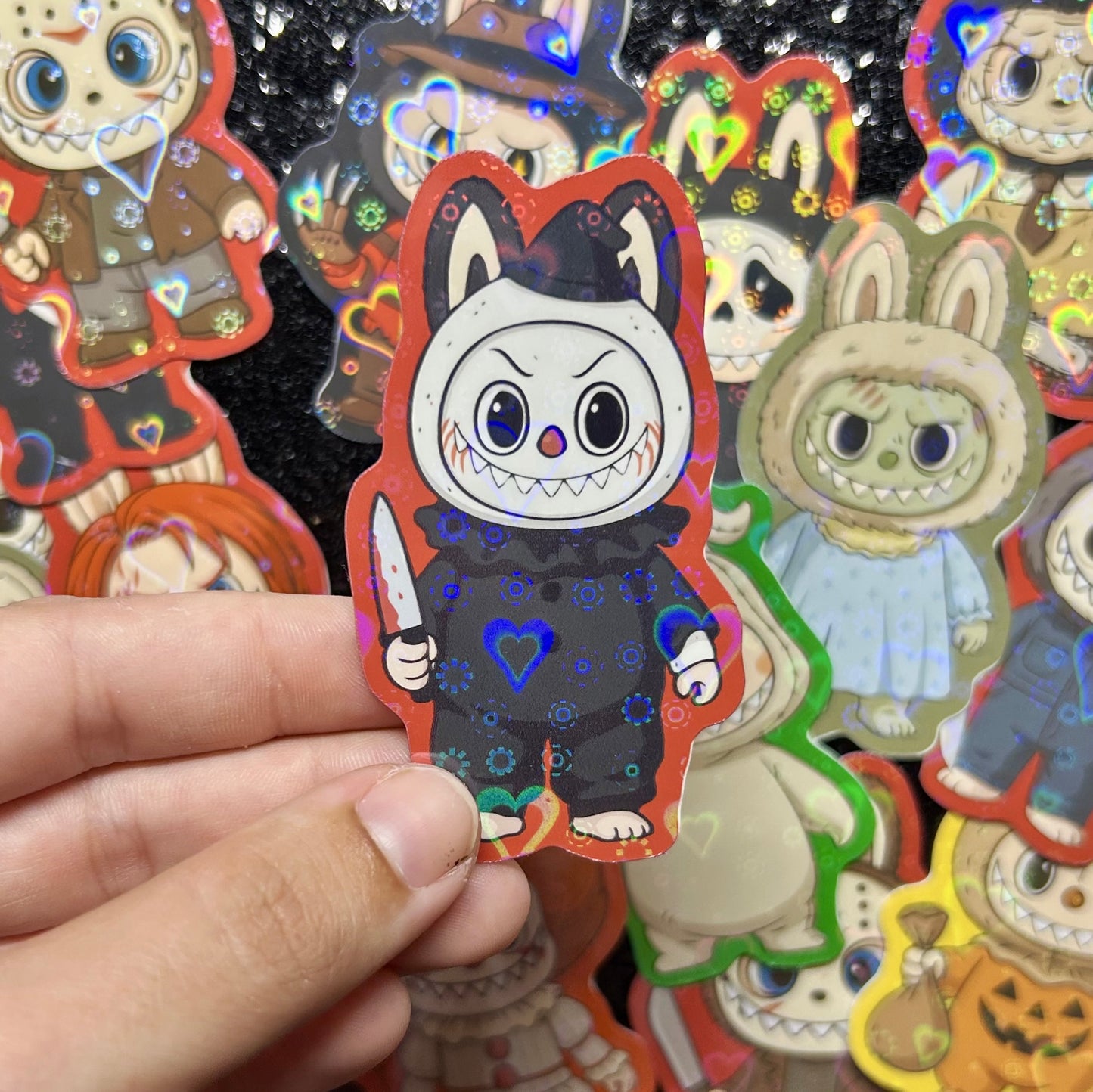 Hand holding a sticker of a Terrifier Art the Clown Labubu sticker, surrounded by other colorful stickers on a black background
