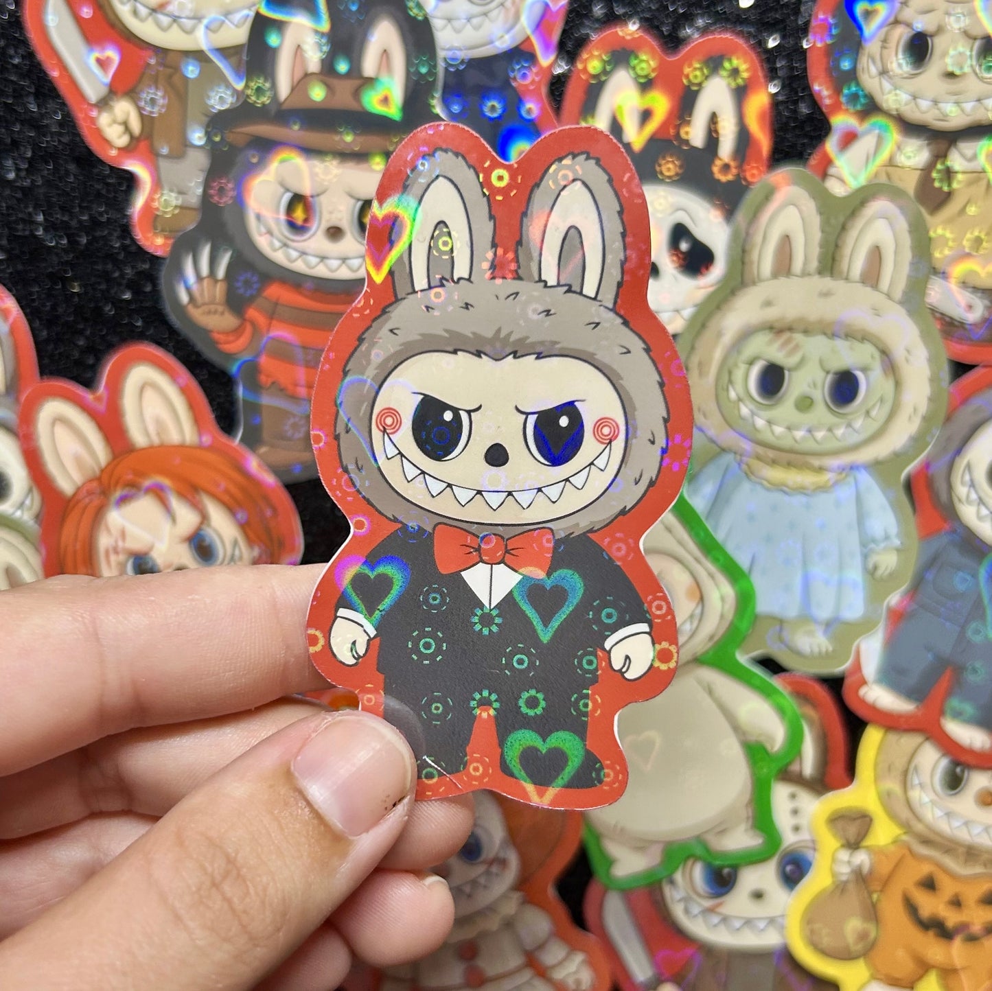 Hand holding a sticker of a SAW Billy Jigsaw Labubu sticker, surrounded by other colorful stickers on a black background