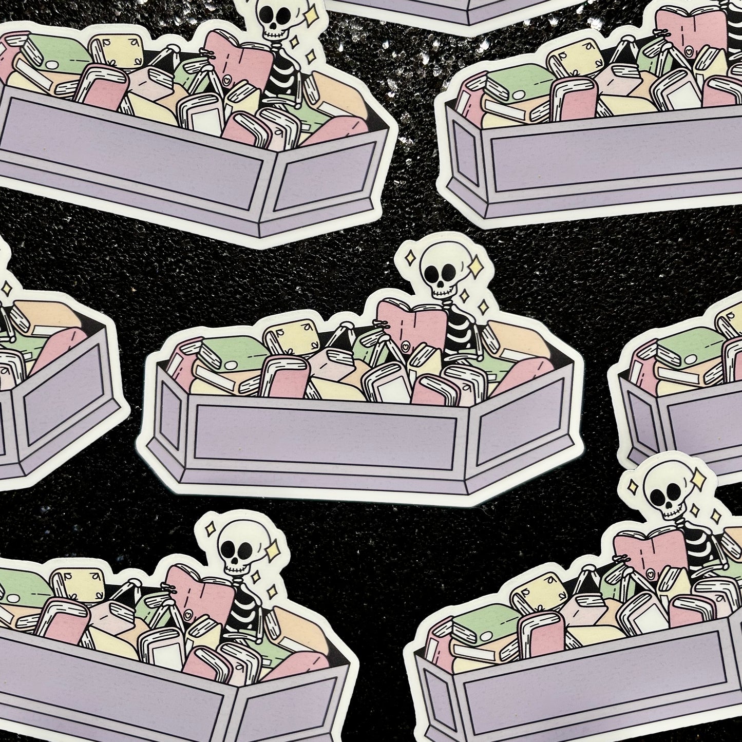 Stickers of a skeleton sitting in a coffin full of books reading a book on a black background