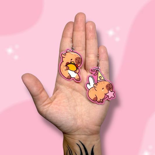 Hand holding Taco capybara and princess capybara earrings on a pink background.