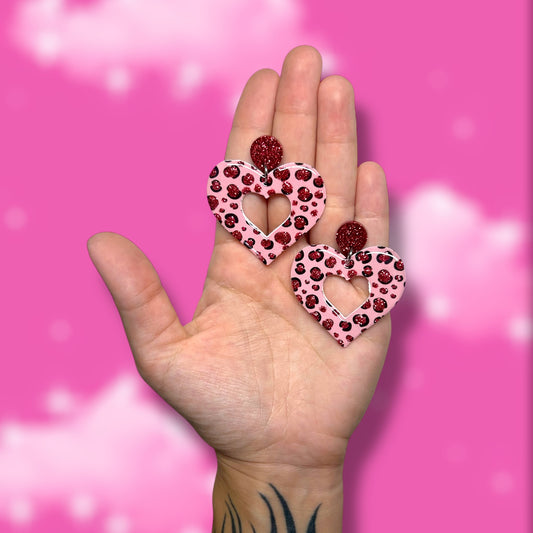 Hand holding glittery pink cheetah-print mod heart-shaped earrings on a blurred pink background.