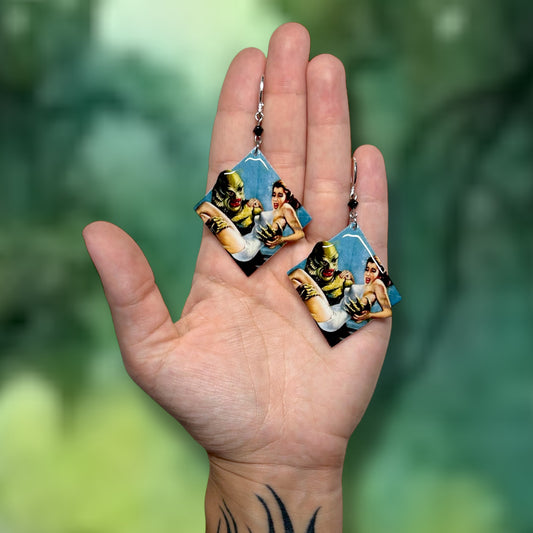 Hand holding Creature from the Black Lagoon earrings on a blurred green background.