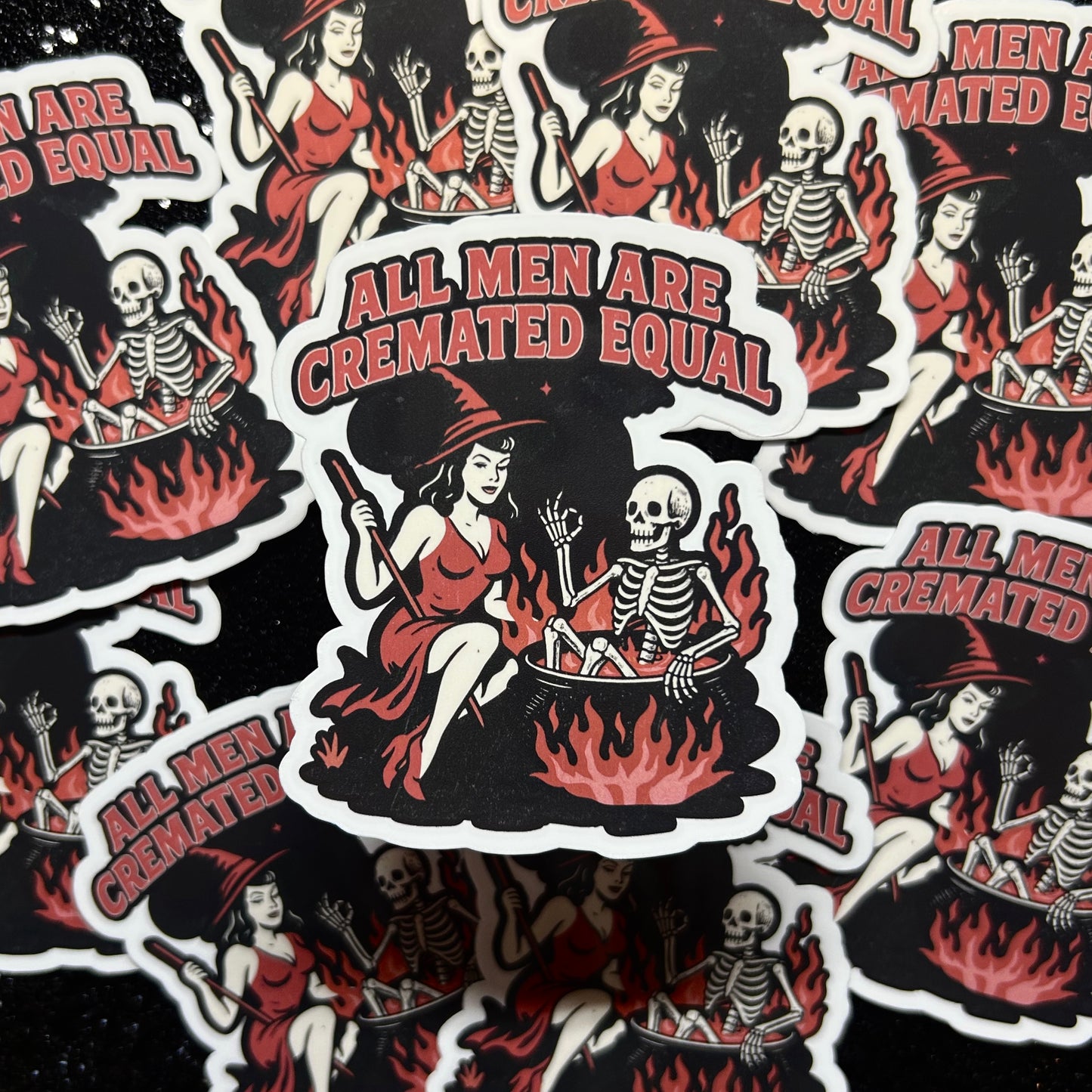 Sticker with skeleton in cauldron over fire and woman dressed like a witch and text 'All Men Are Cremated Equal' on a black background.