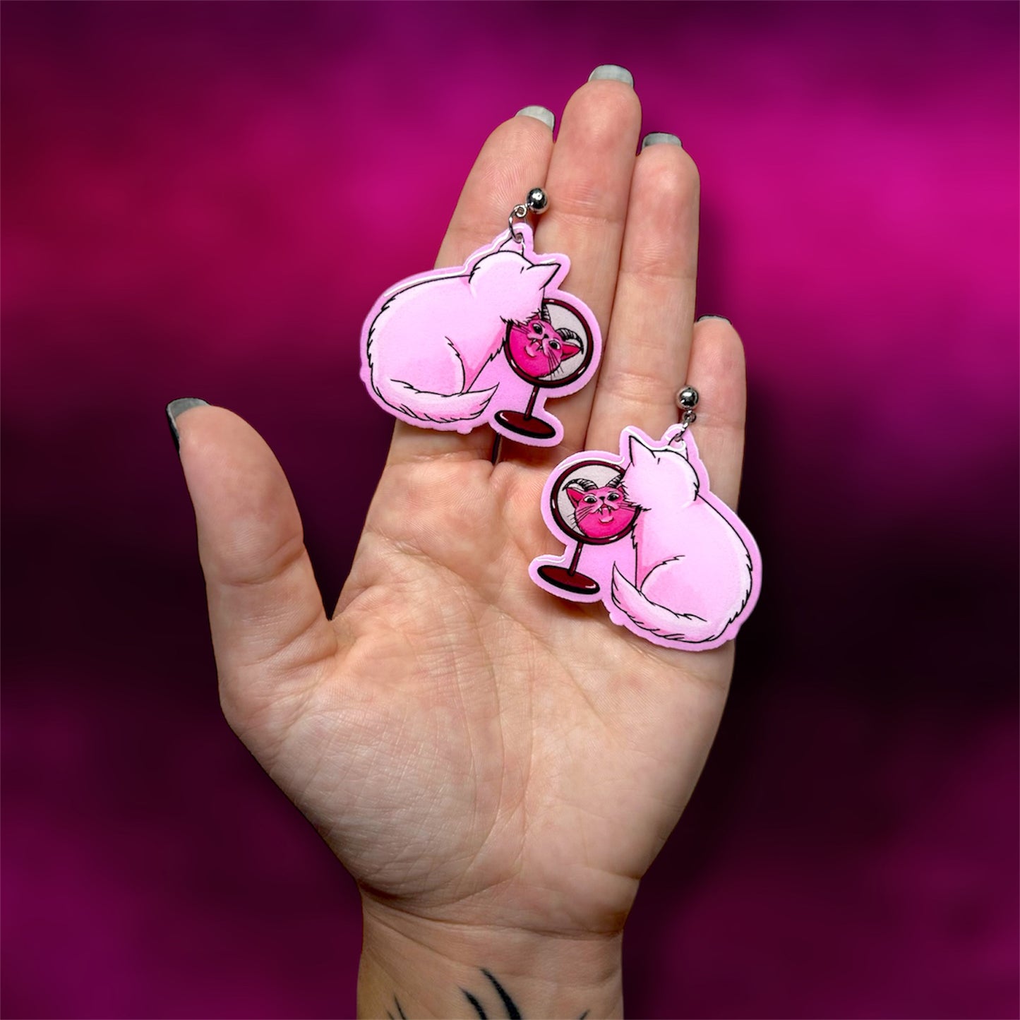 Hand holding a pair of earrings of a pink demon cat looking into a mirror against a pink background