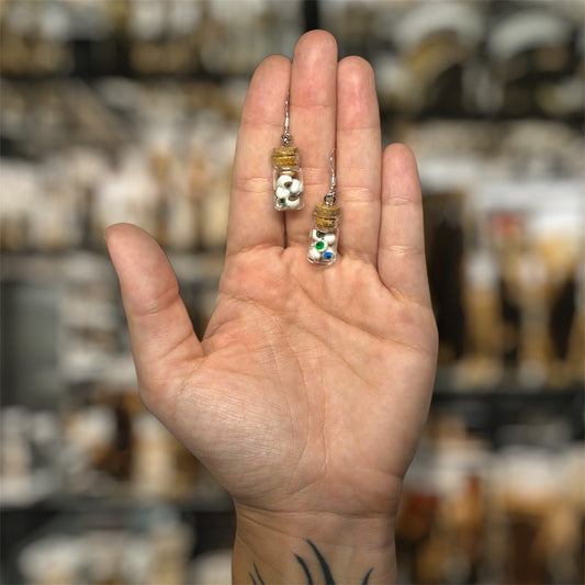 Hand holding small glass jar earrings containing brown eyes, blue eyes, and green eyes in water on a blurred background.