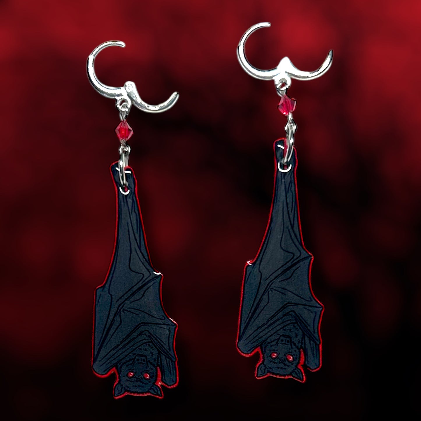 Upside-down hanging vampire bat earrings on a blurred red and black background.