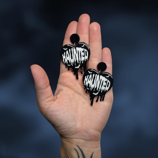Hand holding heart-shaped Halloween earrings with 'HAUNTED' text and bats on a dark background
