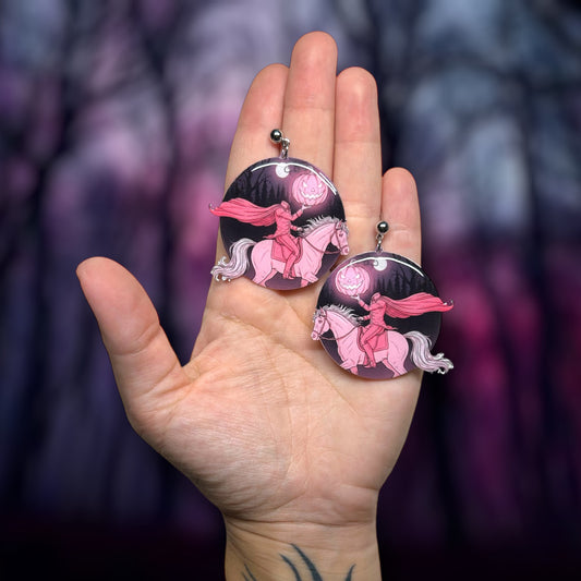 Hand holding pink Halloween headless horseman pink pumpkin jack o lantern earrings on a dark blurry background.