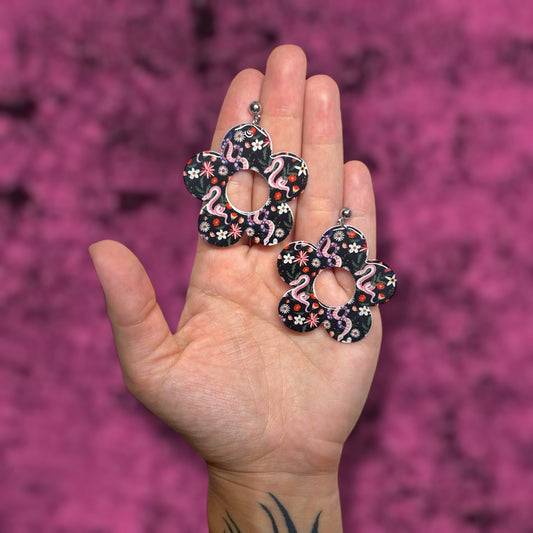 Hand holding floral and snake flower mod earrings on a pink background
