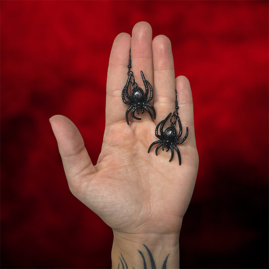 Hand holding black spiders with red eyes earrings on a blurred red and black background.