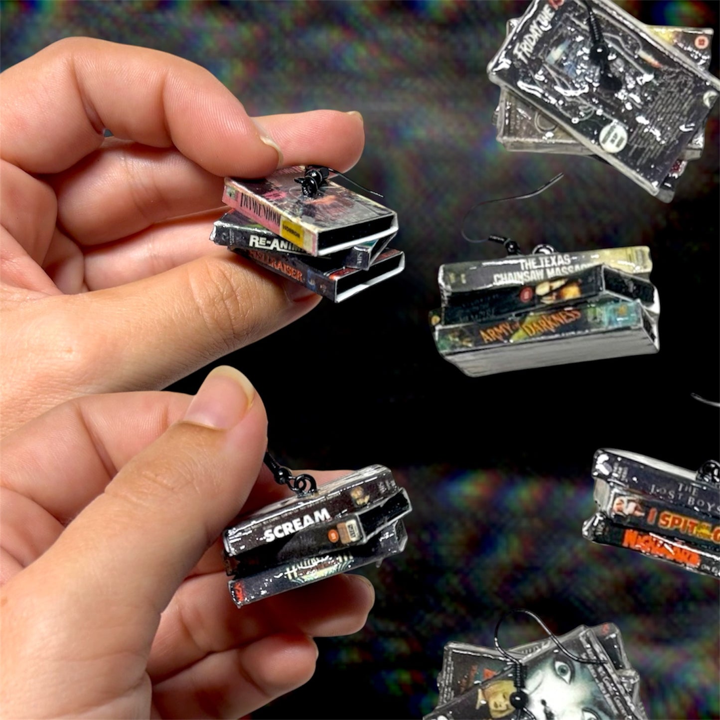 Hands holding stacked VHS tape statement earrings, featuring the movies Scream, Chopping Mall, Halloween 3, Frankenhooker, Re-Animator, and Hellraiser, shown against a dark, grainy TV static background, and surrounded by other stacked VHS dangle earrings.