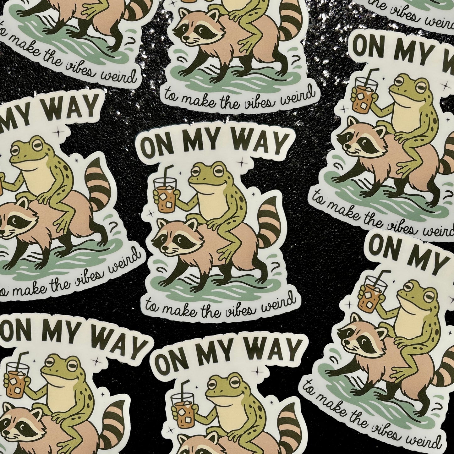 Stickers of a frog drinking iced coffee riding a raccoon with text 'On my Way to Make the Vibes Weird' on a black background