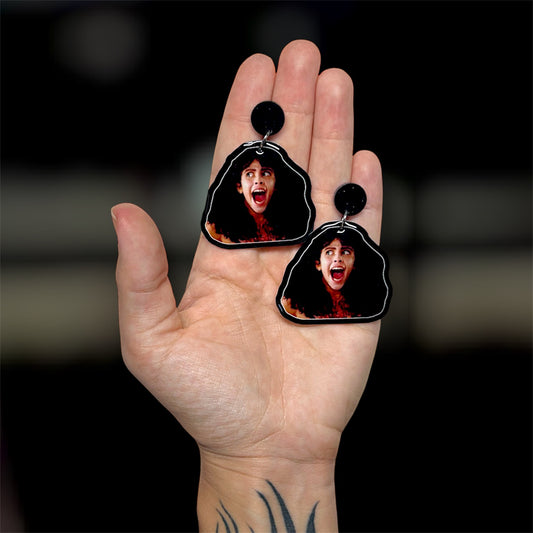 Hand holding a pair Sleepaway Camp bloody Angela screaming resin horror dangle earrings on a blurry background