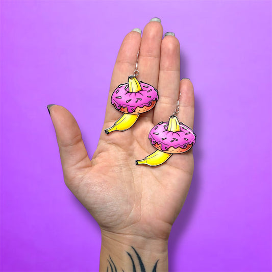 Hand holding a pair of bananas inside pink sprinkle donuts resin dangle earrings on a purple background