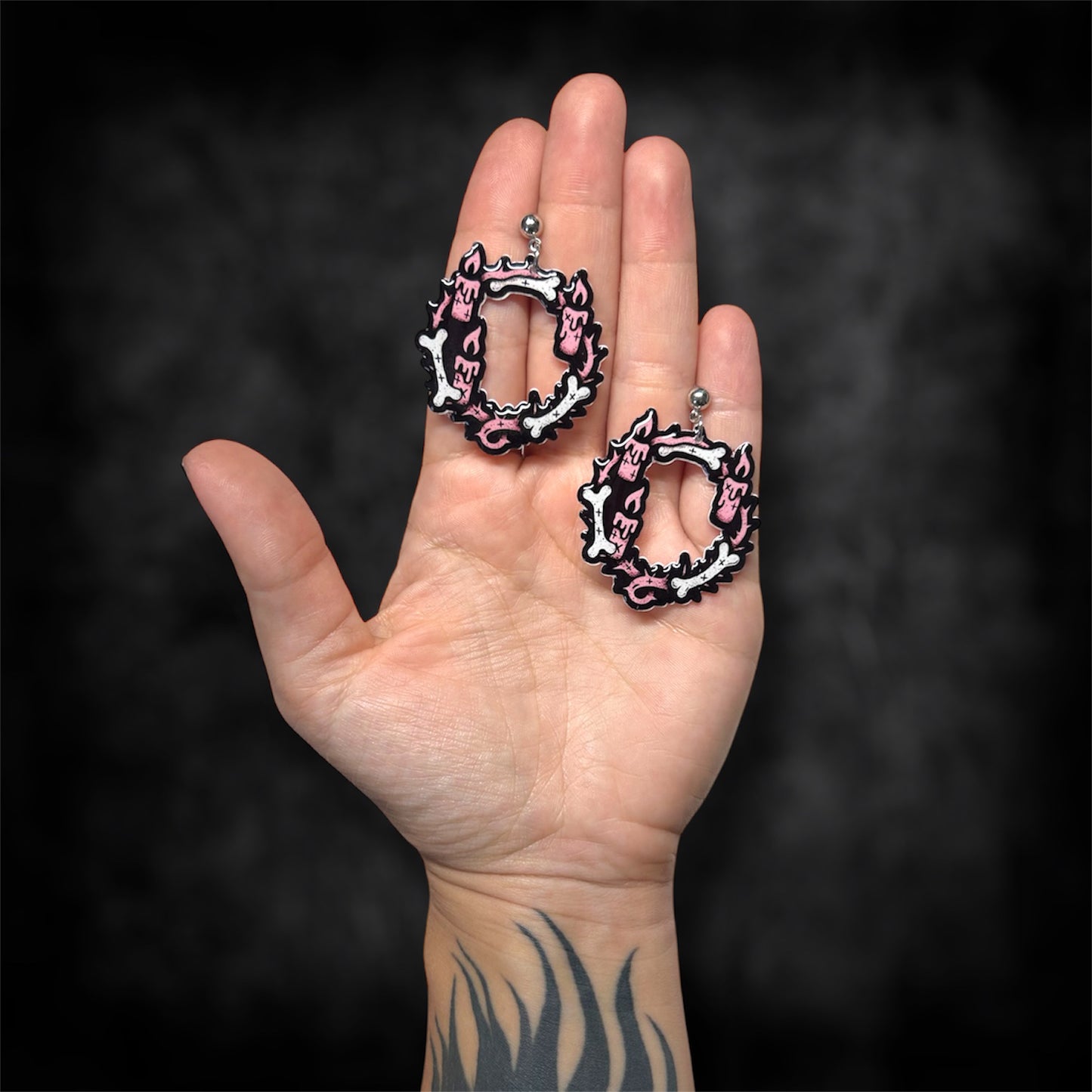 Hand holding pink Satanic candles and bones hoop earrings on a blurry black background