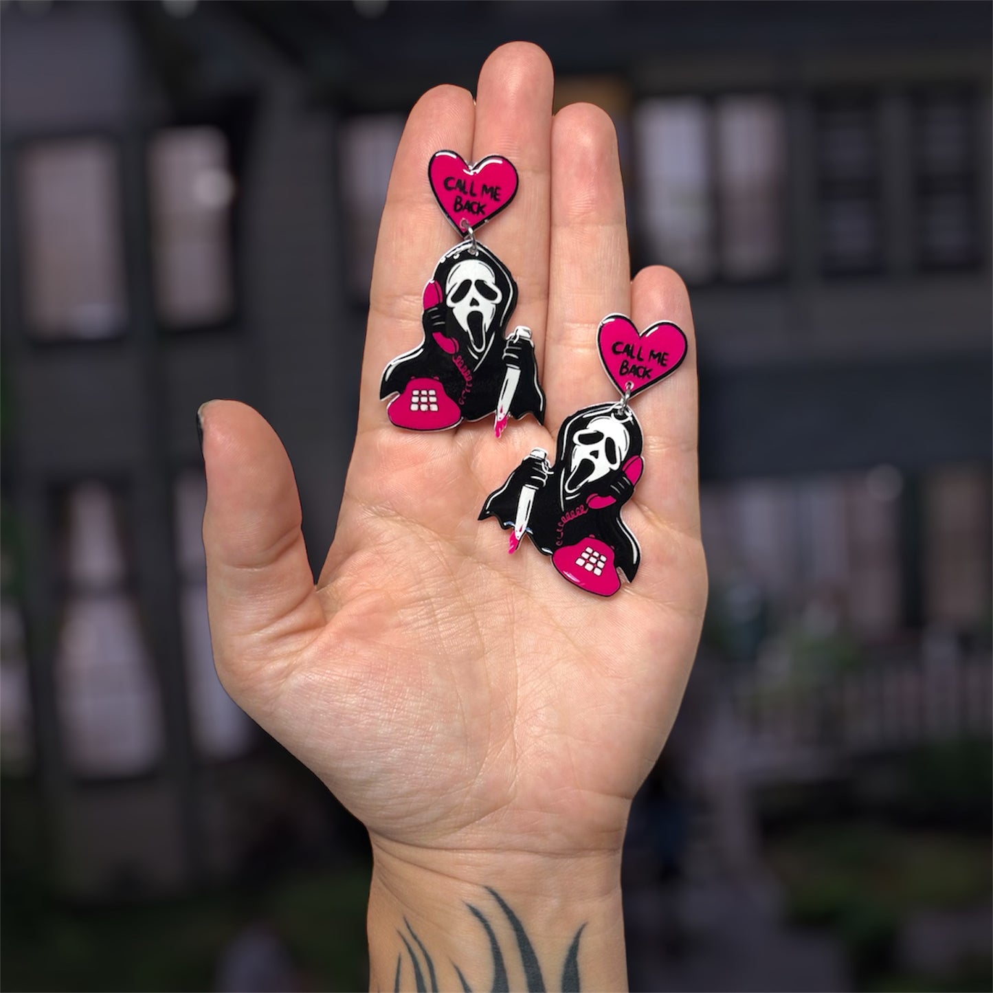Hand holding pink, white, and black Scream Ghostface "Call Me Back" resin dangle earrings, with Ghostface holding a bloody knife and a pink corded phone