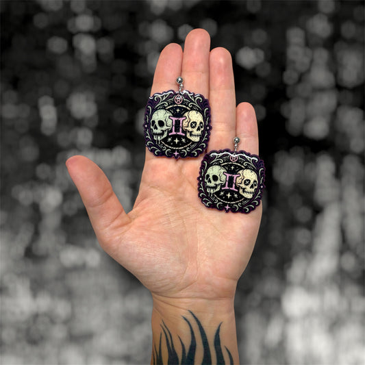 Hand holding a pair of Gemini zodiac sign twin skulls witchy resin dangle earrings on a blurry black and white background