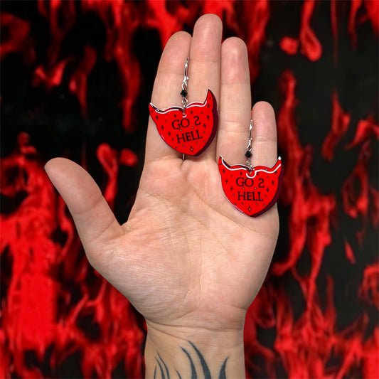 Hand holding red and black sparkly devil horns heart "Go 2 Hell" beaded resin dangle earrings on a blurry fiery background
