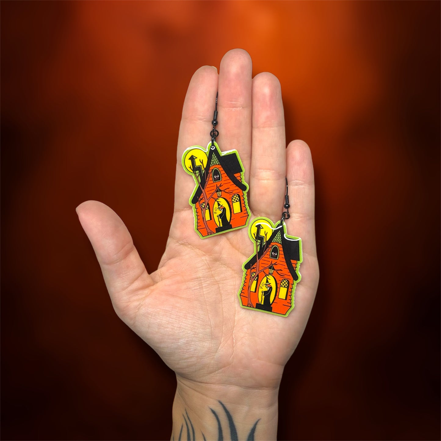 Hand holding Halloween spooky haunted house with witch earrings on an orange background
