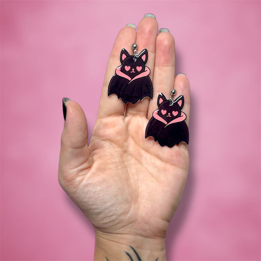 Hand holding a pair of bat earrings with pink heart eyes on a pink background