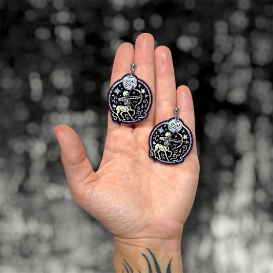 Hand holding a pair of Sagittarius zodiac sign centaur with bow and arrow resin dangle earrings on a blurry black and white background