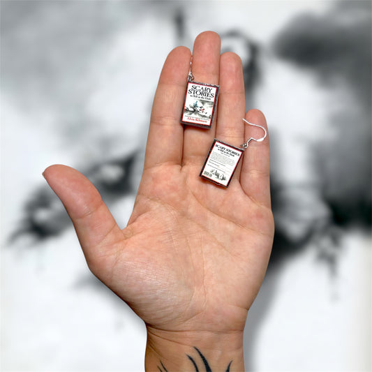 Hand holding a pair of Scary Stories to Tell in the Dark nostalgic horror poetry miniature book earrings on a blurry black and white background