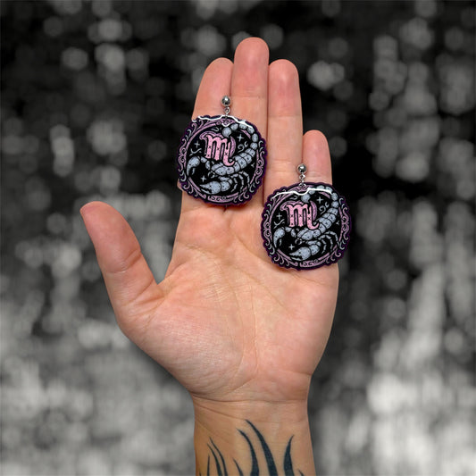 Hand holding a pair of Scorpio zodiac sign scorpion resin dangle earrings on a blurry black and white background