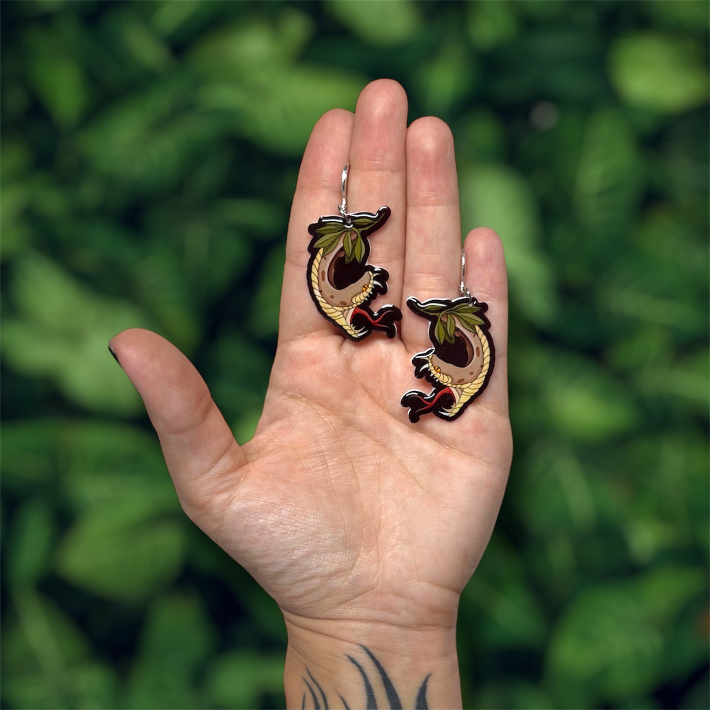 Hand holding spooky garden hissing snake cottagecore leaf resin dangle earrings on a green leafy blurred background