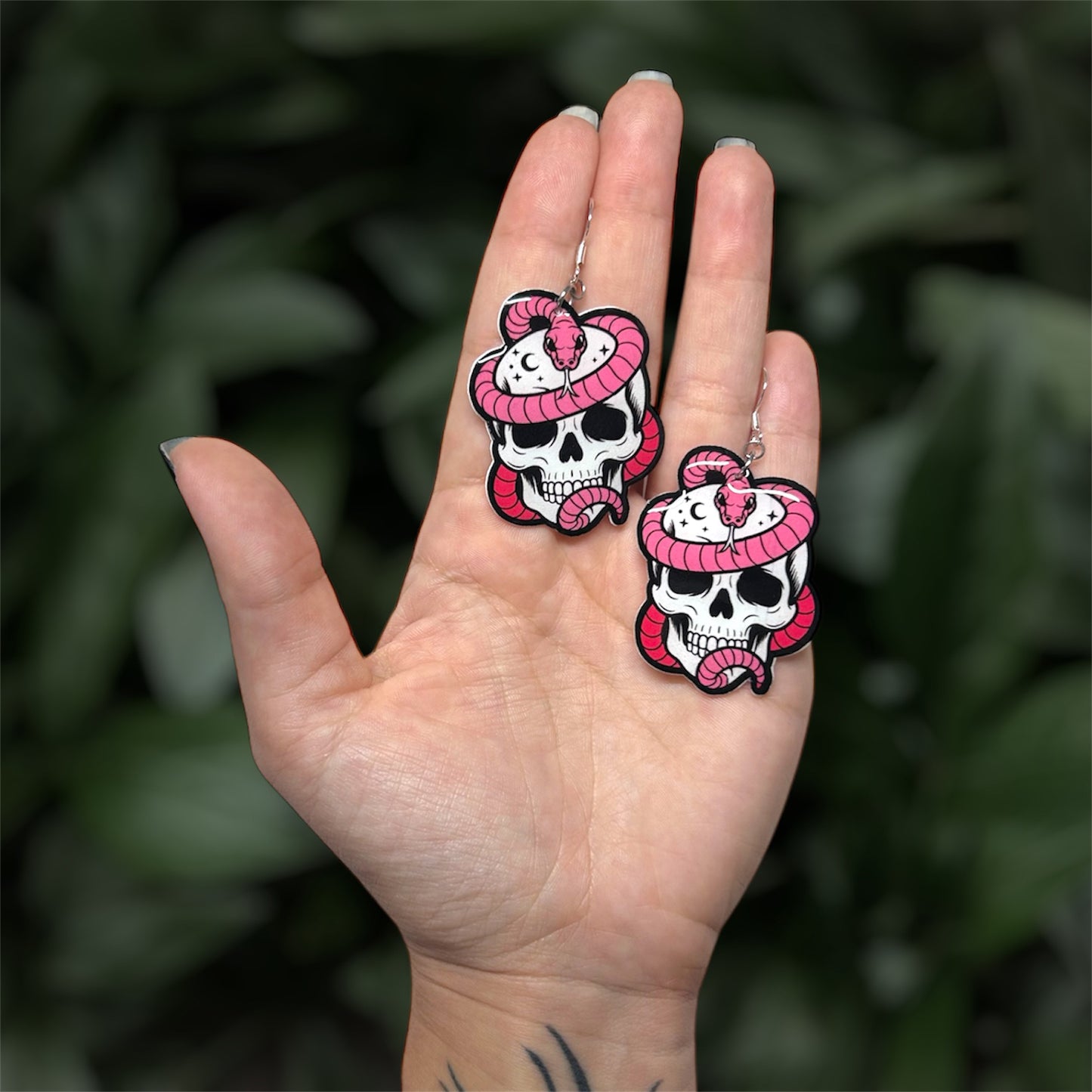hand holding resin dangle earrings of a pink snake wrapped around a skull with stars and moons, on a blurry green background