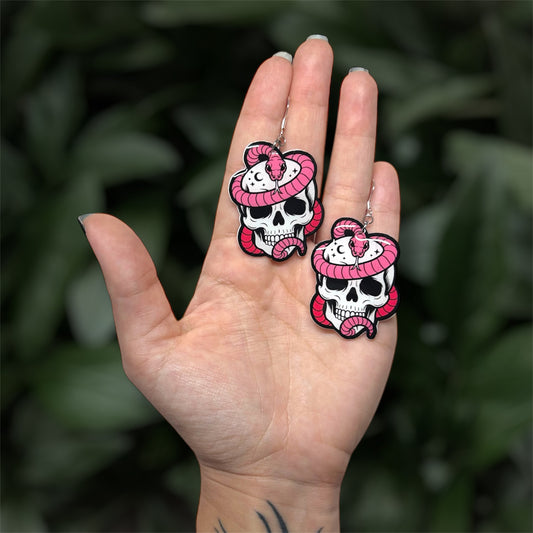 hand holding resin dangle earrings of a pink snake wrapped around a skull with stars and moons, on a blurry green background