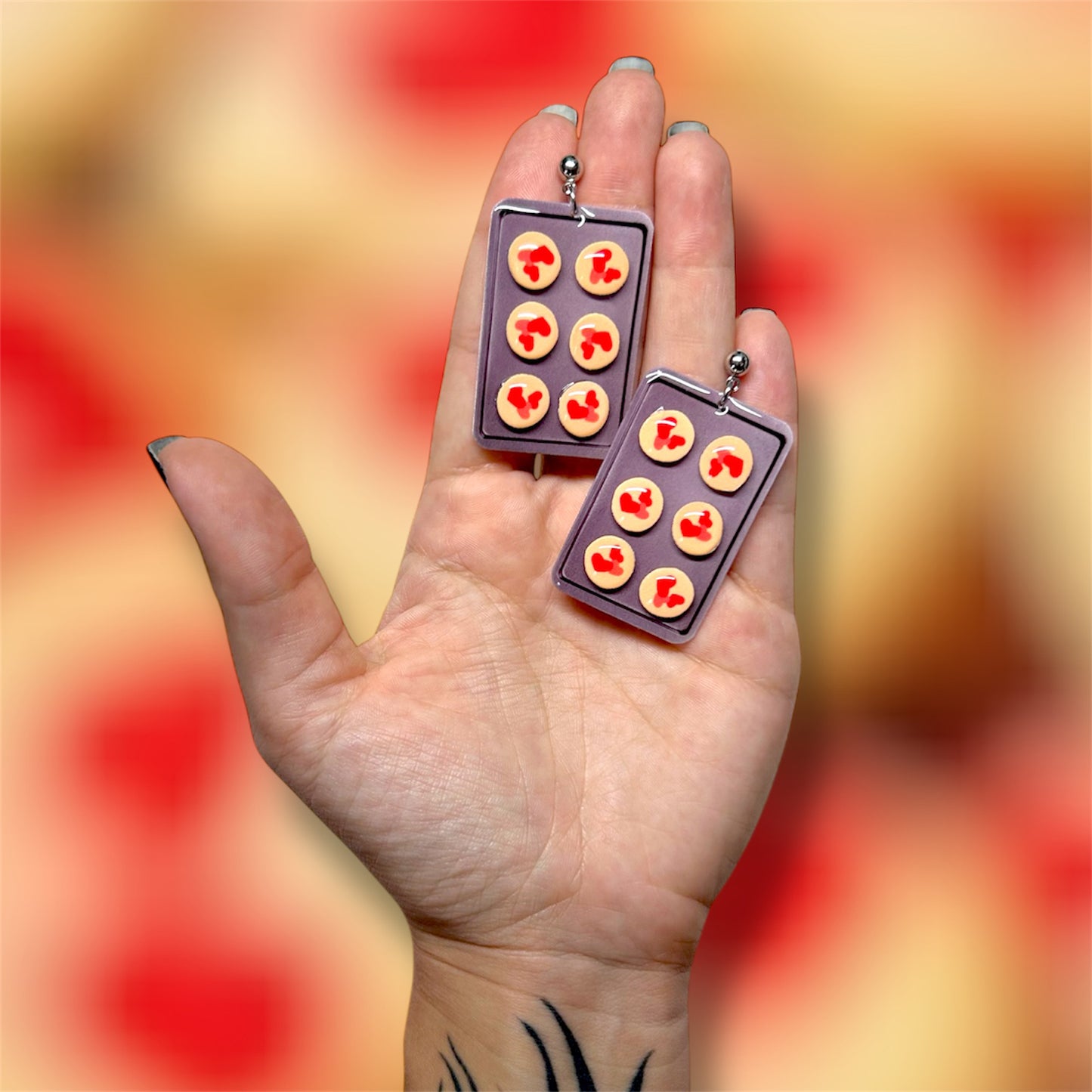 Hand holding a pair of earrings shaped like a cookie tray with red and pink heart Pillsbury Valentine's Day cookies on a colorful background