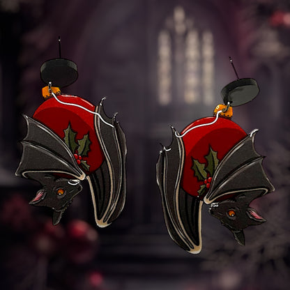 A pair of earrings designed as stylized bats with red and green Christmas-themed details, hanging from hypoallergenic stainless steel studs.
