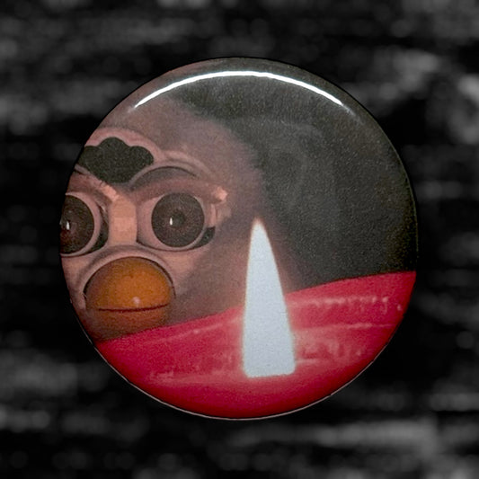 Pinback button of a Furby staring into a fire on a blurry black and white background.
