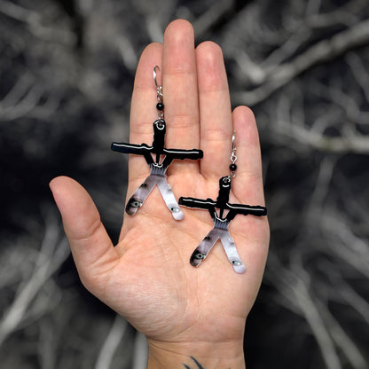 Hand holding Blair Witch Project stick figure earrings depicting Heather's confessional on a blurred forest background.