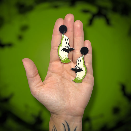 Hand holding glowing Halloween blow mold ghost holding a bat earrings on a blurred green background.