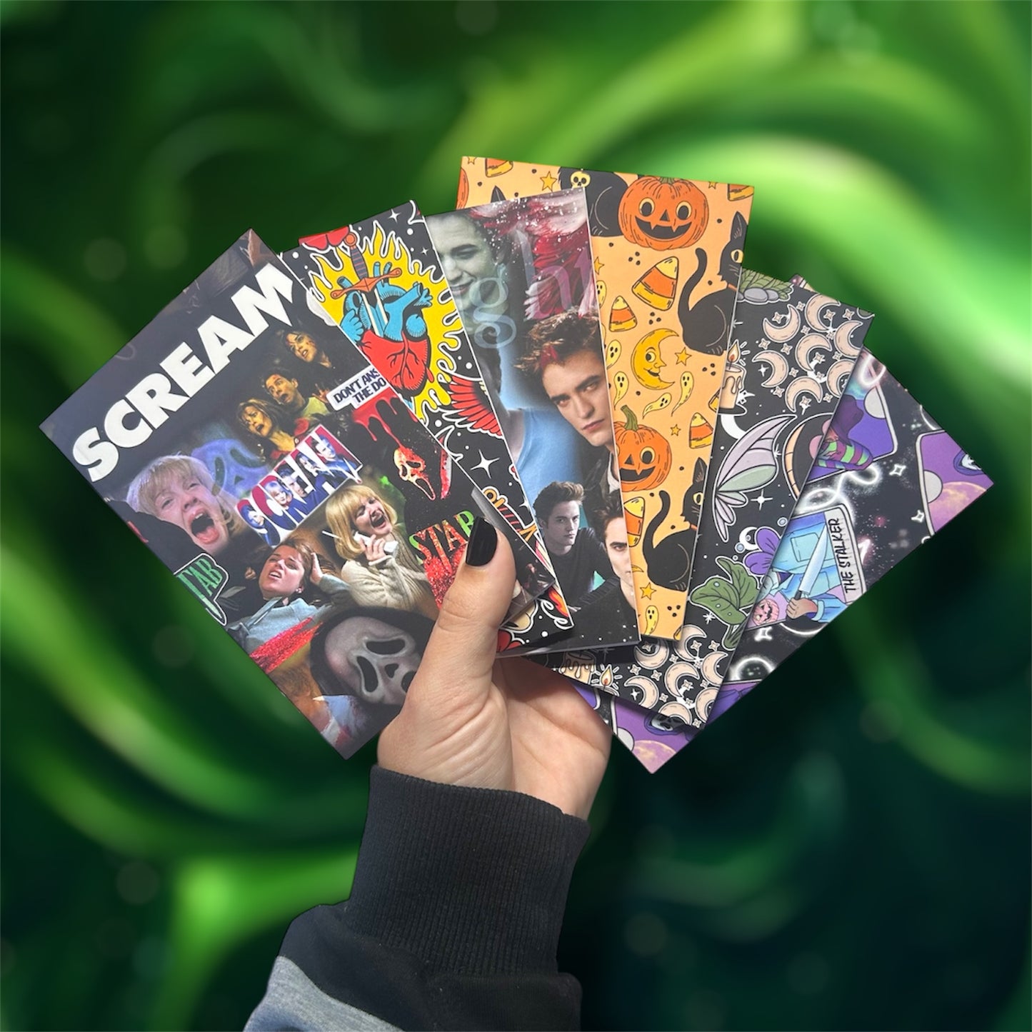 Hand holding several pocket notebooks, including Scream, traditional tattoo art, Twilight, Halloween, cottagecore, and horror tarot, on a blurred green background