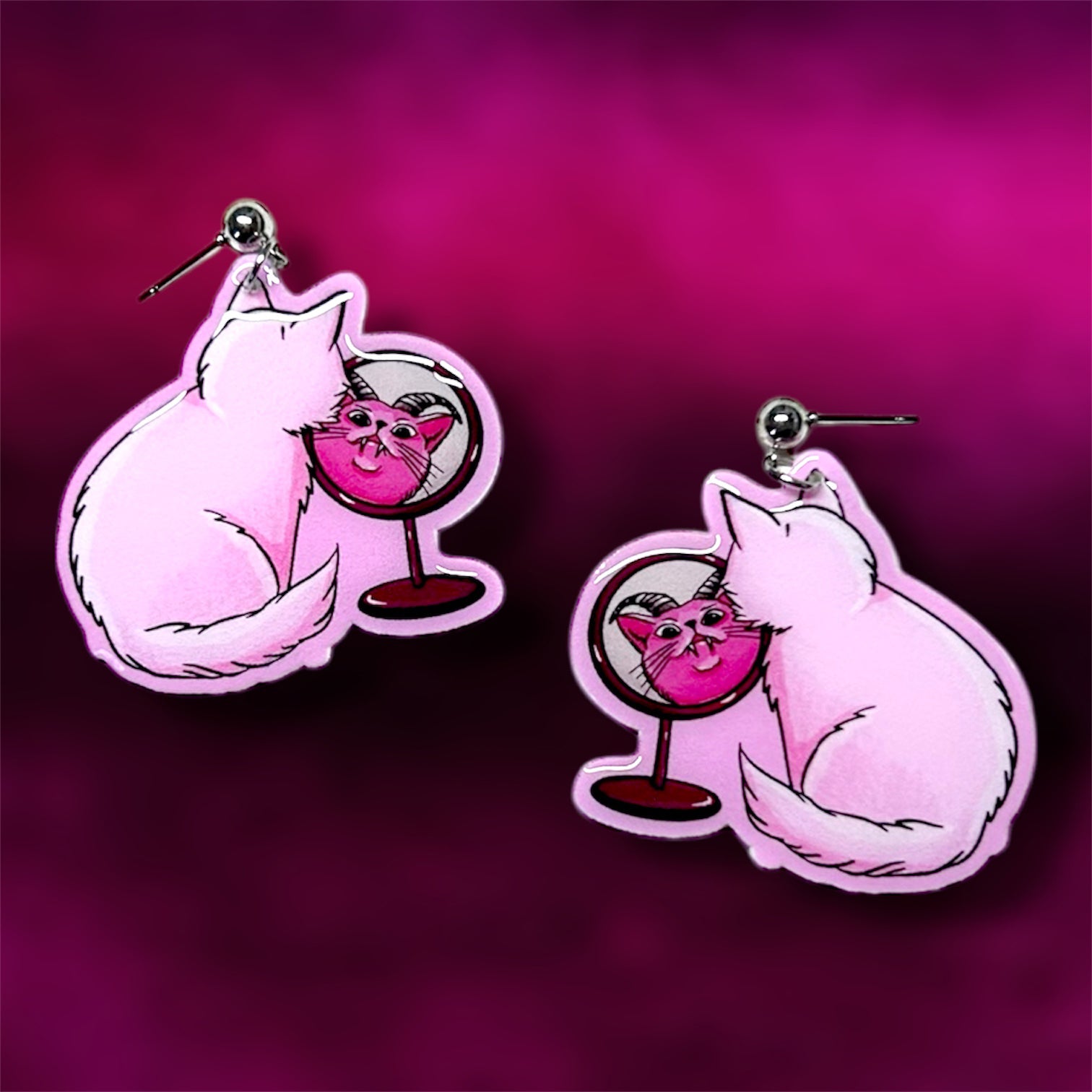 A pair of earrings of a pink demon cat looking into a mirror against a pink background