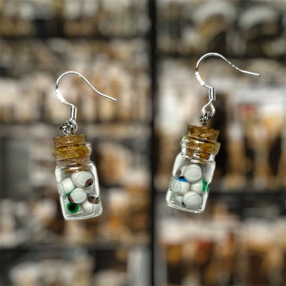 Small glass jar earrings containing brown eyes, blue eyes, and green eyes in water on a blurred background.
