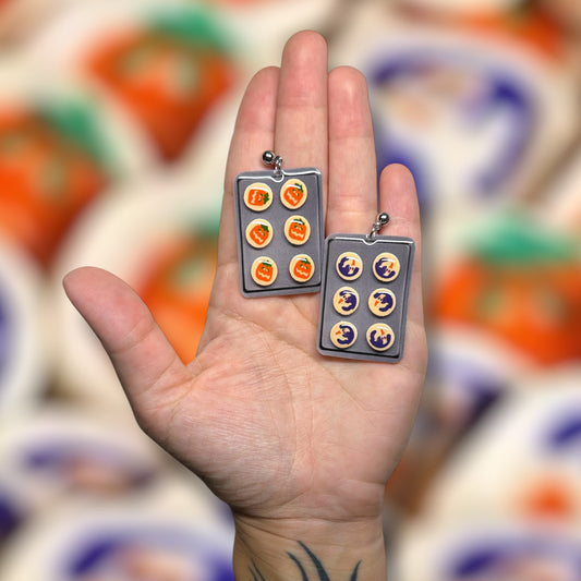 Pillsbury ghost cookie Pillsbury pumpkin cookie Halloween cookie sheet spooky cute resin stud earrings being held in a hand on a blurry orange and purple background