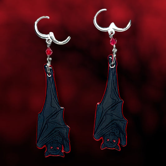 Upside-down hanging vampire bat earrings on a blurred red and black background.