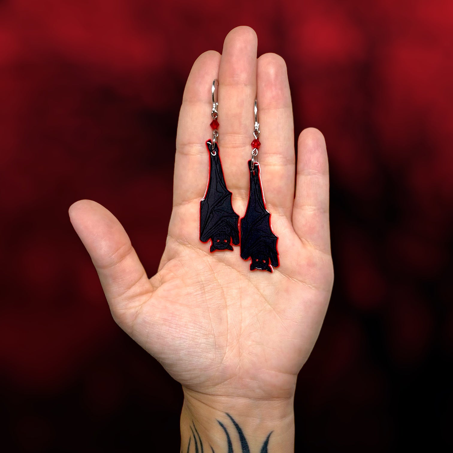 Hand holding upside-down hanging vampire bat earrings on a blurred red and black background.