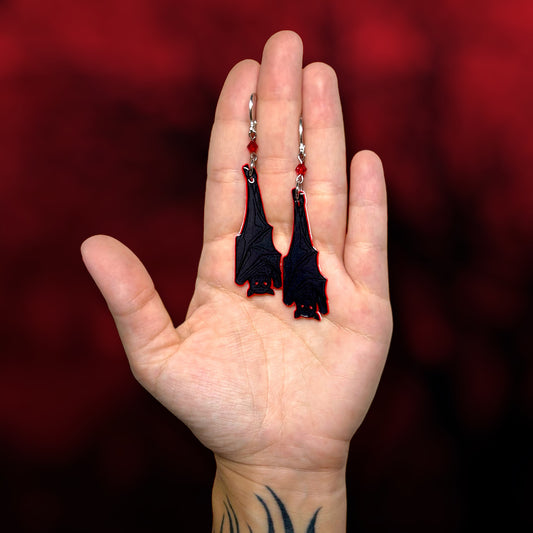 Hand holding upside-down hanging vampire bat earrings on a blurred red and black background.