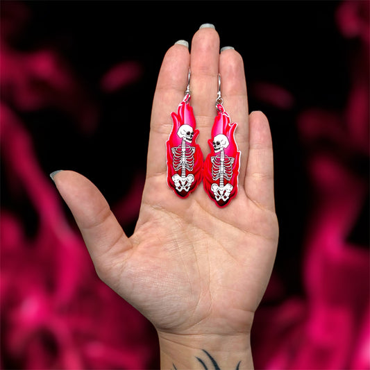 Hand holding a pair of skeleton in pink fire resin statement earrings on a blurred pink and black background