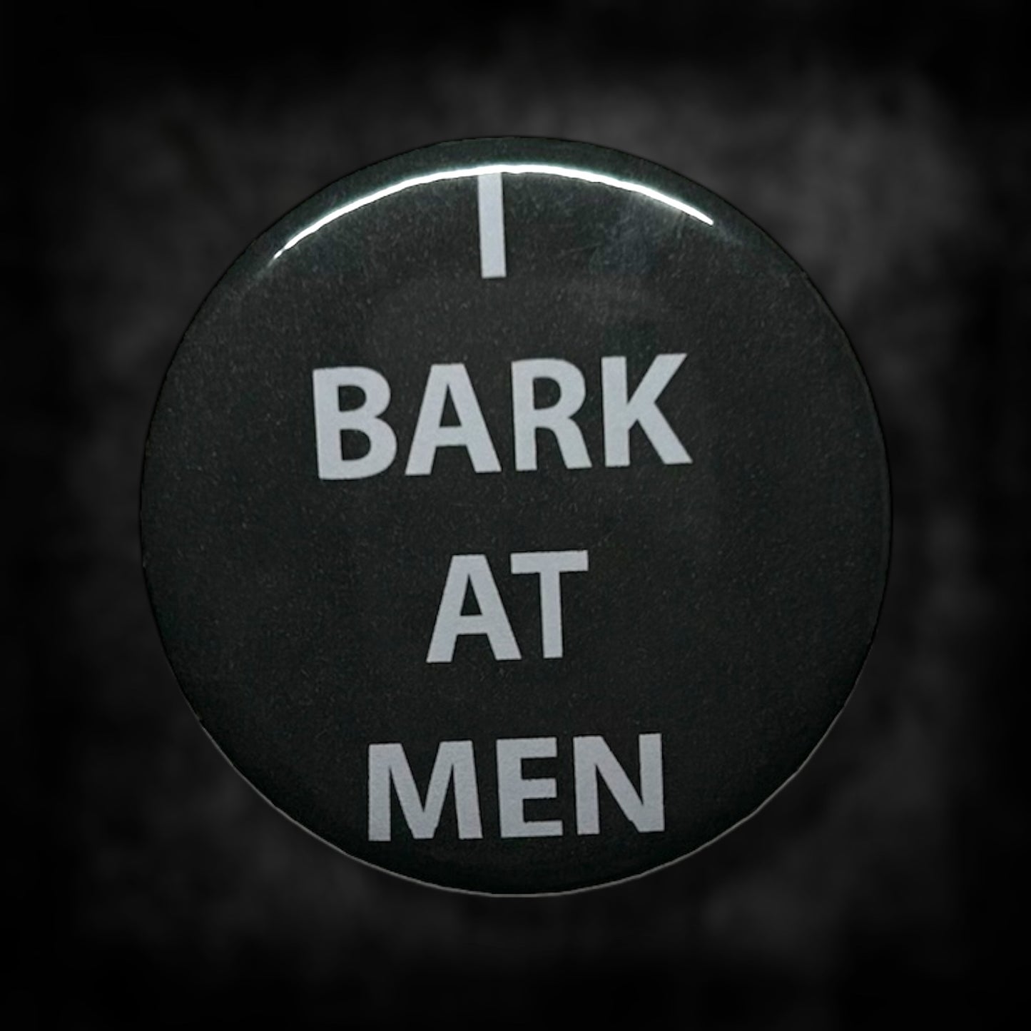 Black pinback button with white text that read "I BARK AT MEN" on a blurred black and white background.