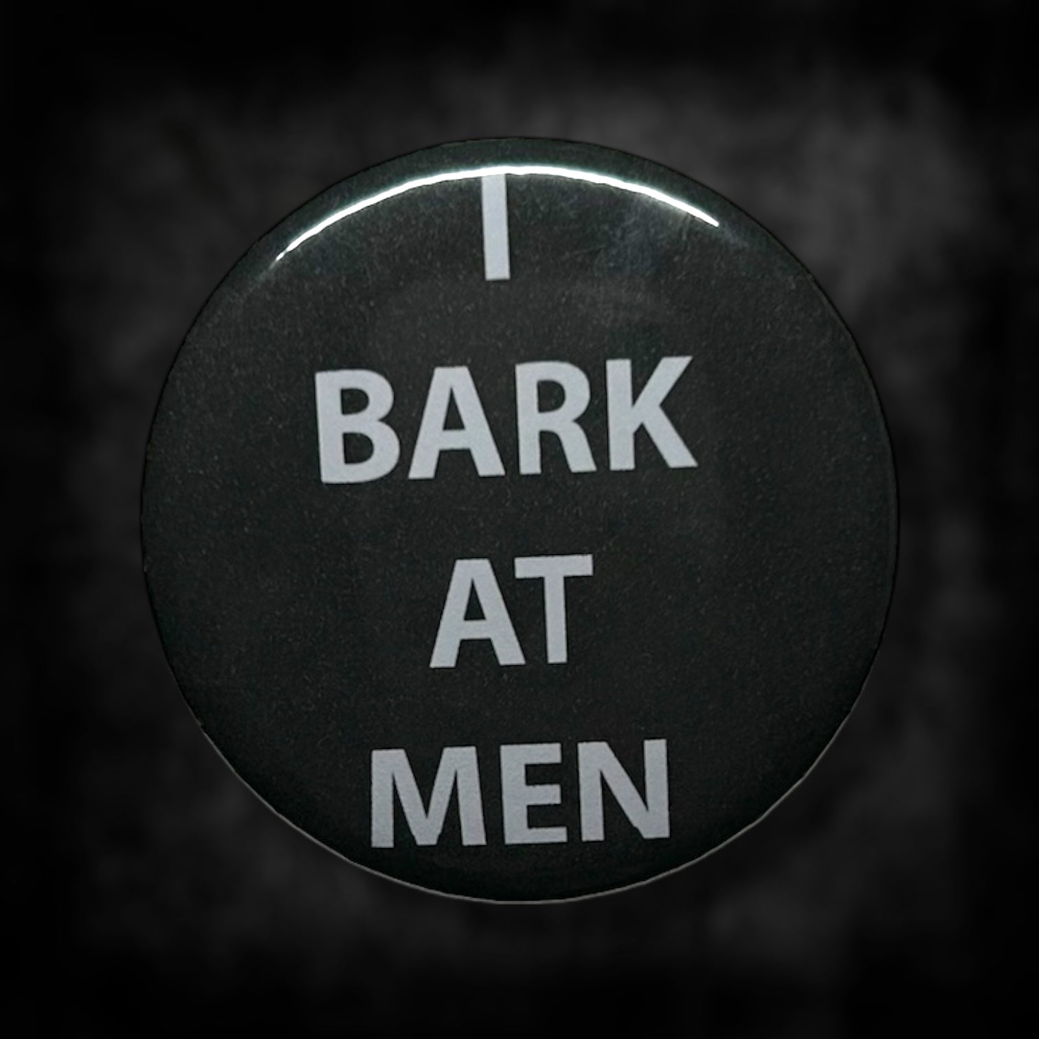 Black pinback button with white text that read "I BARK AT MEN" on a blurred black and white background.