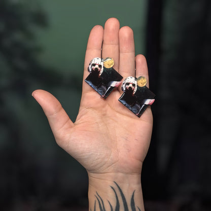Hand holding a pair of Friday the 13th Jason Voorhees hocket mask with bloody axe resin statement earrings on a blurry green and black background