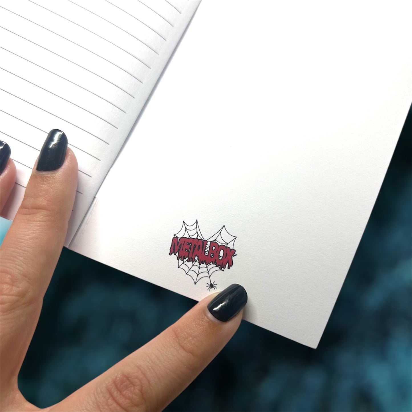 Hand holding an open pocket notebook with the Metalbox logo on the inside back cover on a blurred blue background