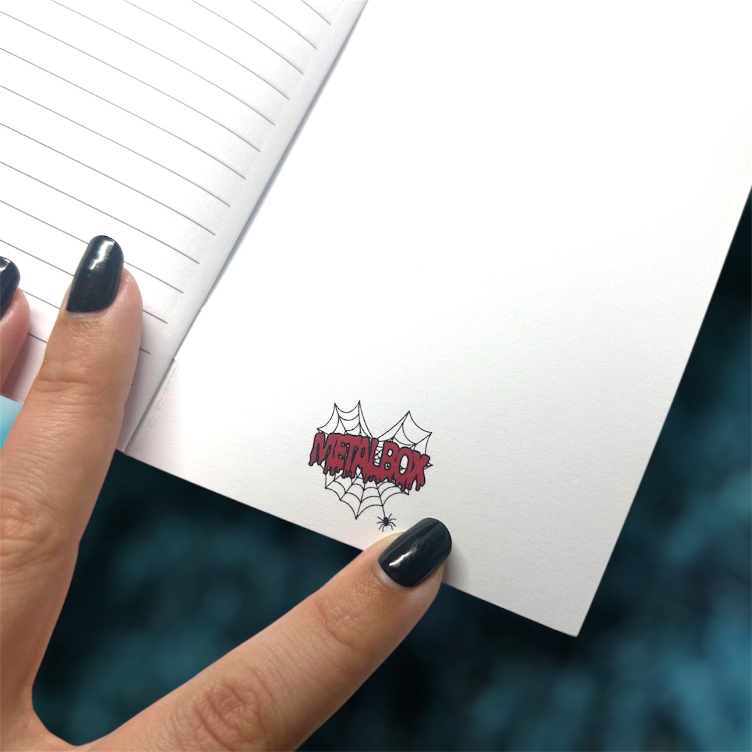 Hand holding an open pocket notebook with the Metalbox logo on the inside back cover on a blurred blue background