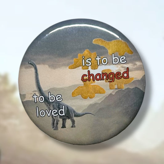 Pinback button of a mountain landscape, a brachiosaurus dinosaur, and dinosaur chicken nuggets with white and red text that reads "to be loved is to be changed" on a blurred bright sunny background.