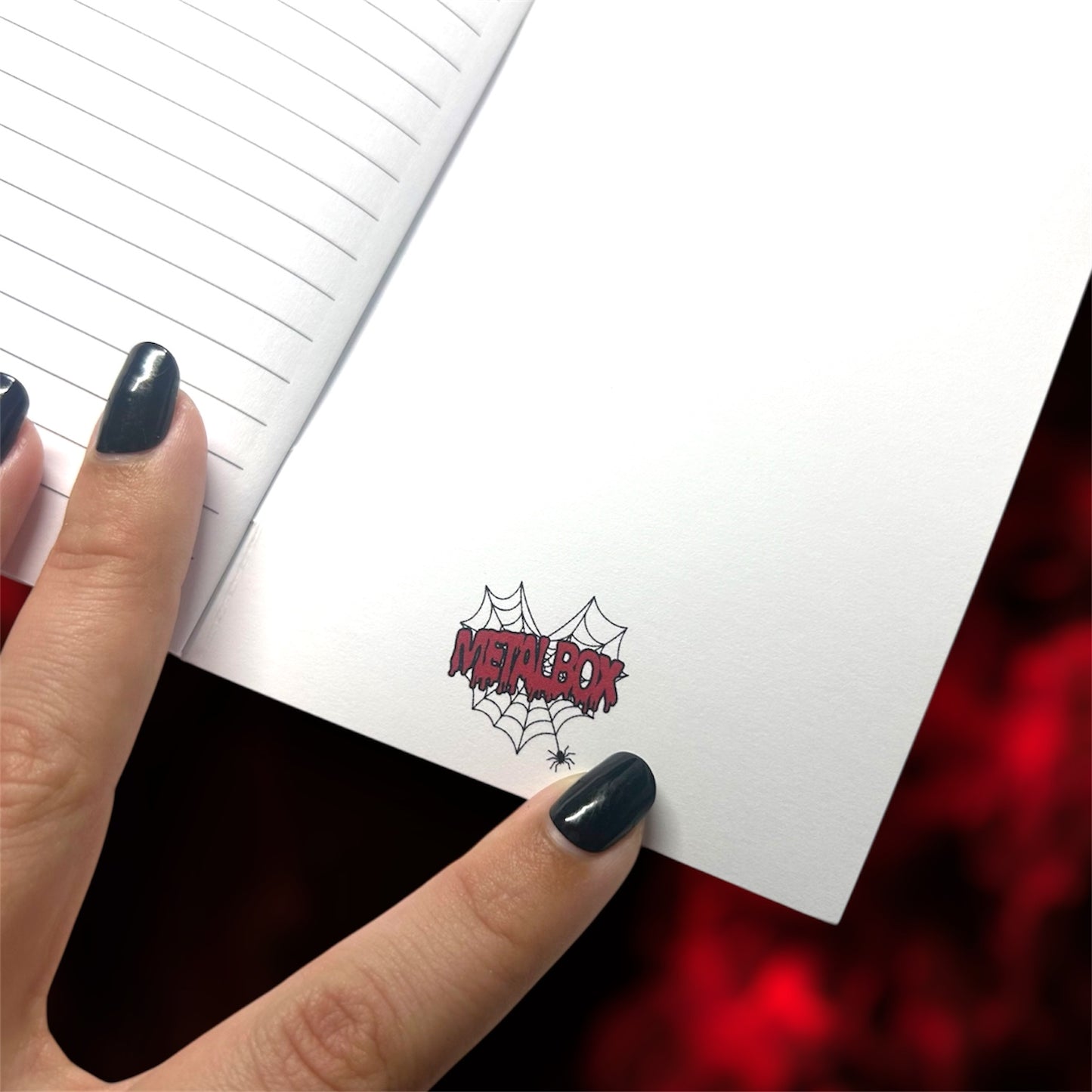 Hand holding an open pocket notebook with the Metalbox logo on the inside back cover on a blurred red background